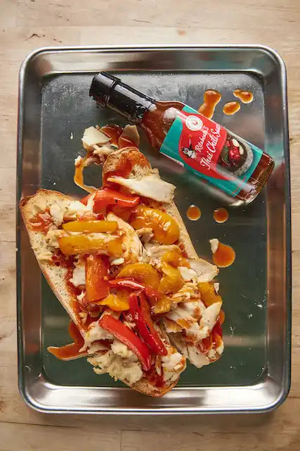 Open-faced sandwich topped with shredded chicken, roasted peppers, and hot sauce alongside a bottle of sauce.