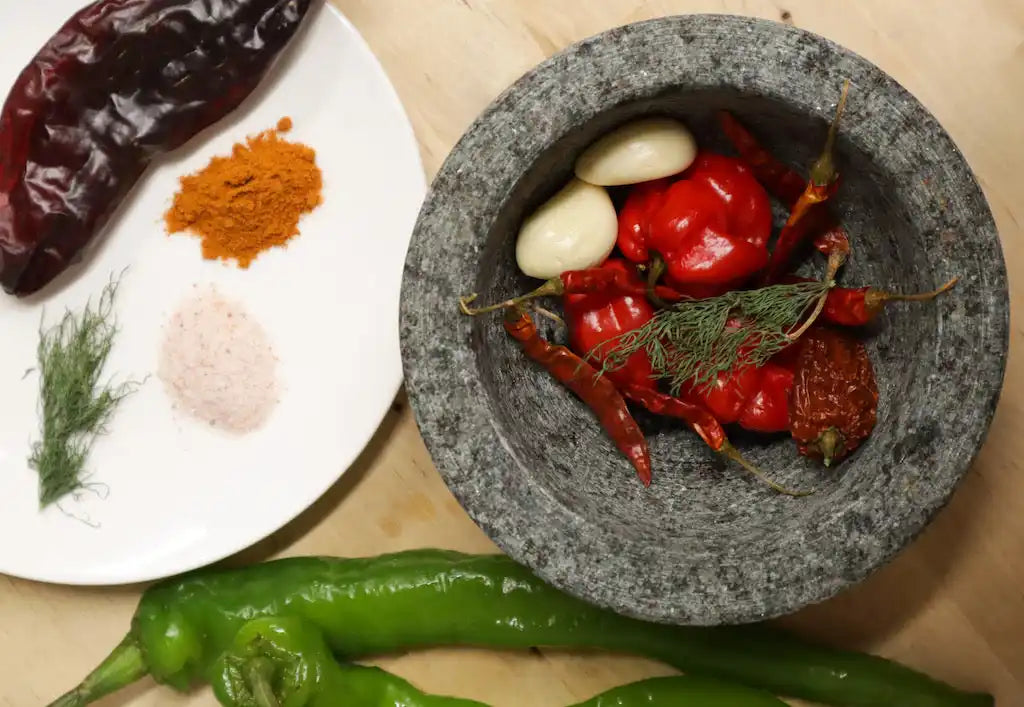 Stone mortar and pestle containing red peppers, garlic cloves, and herbs.