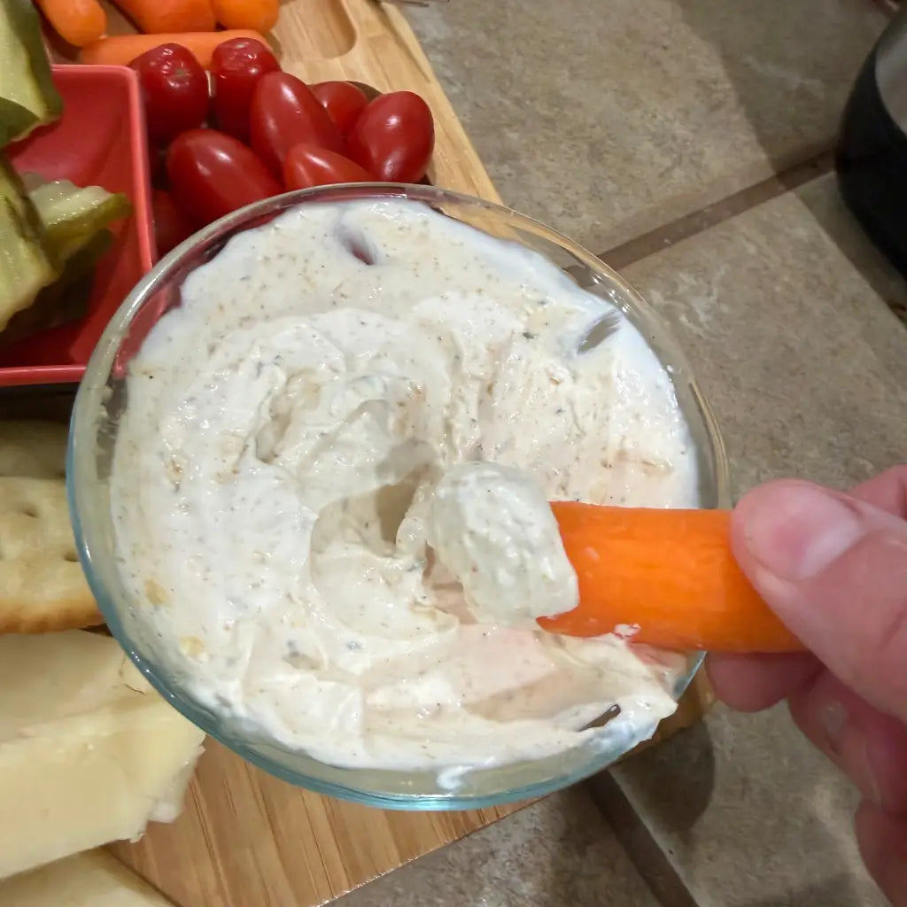 Person dipping a carrot into a bowl of onion dip with a charcuterie board in the background.