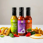 Three hot sauce bottles with colorful labels on a white surface with food items.