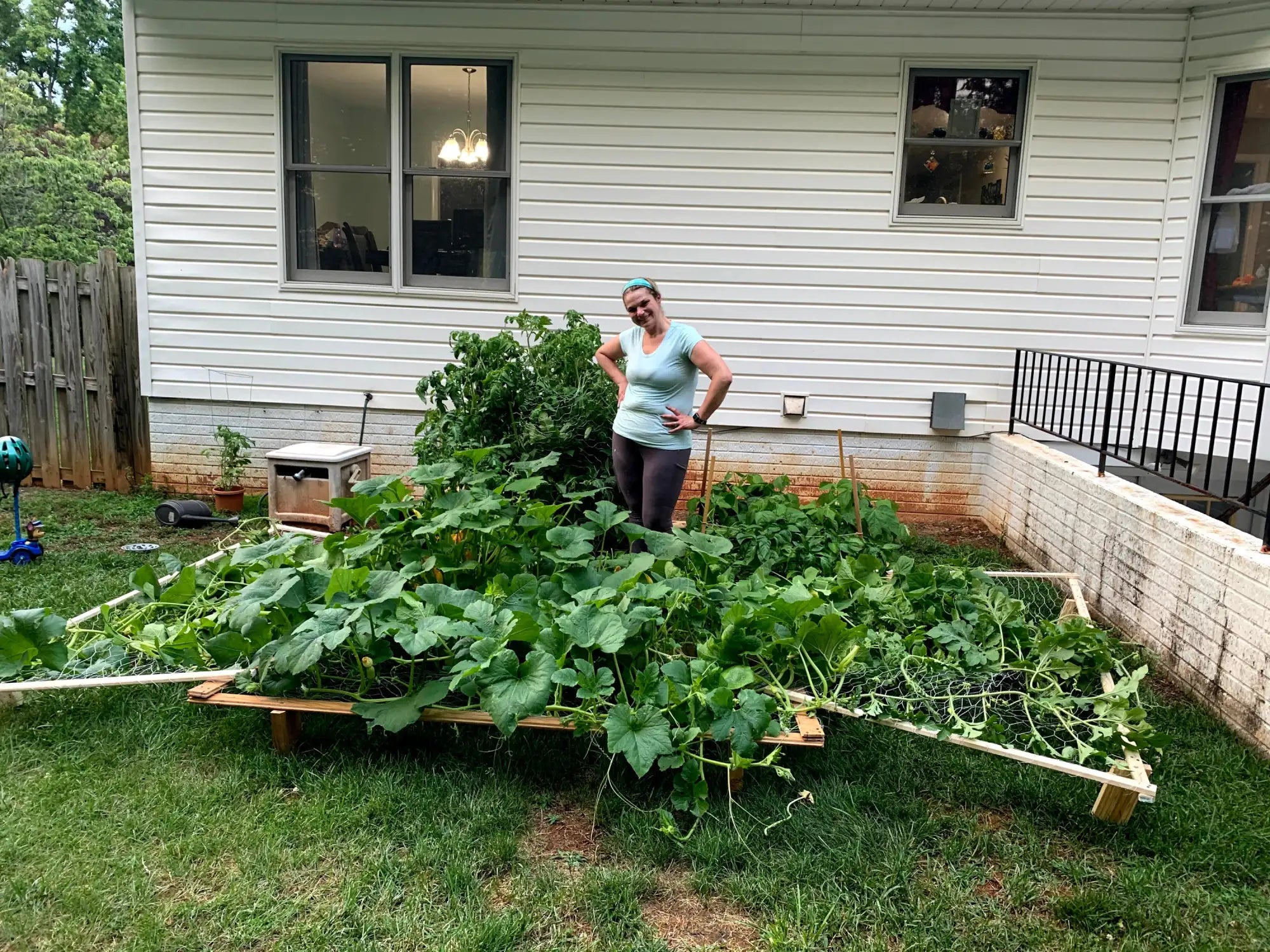 A woman in a light blue top and dark pants stands with hands on hips beside a raised garden bed filled with lush green squash plants.